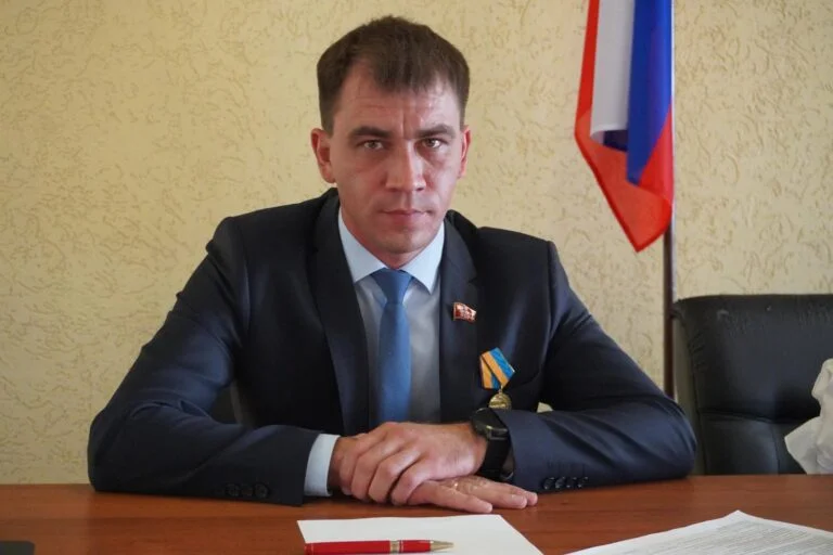 Чоловік у костюмі сидить за столом біля російського прапора