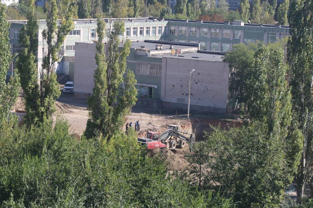Екскаватор працює біля багатоповерхової будівлі серед дерев